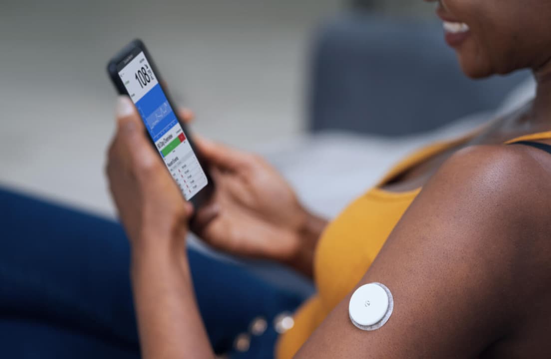 Close-up of a person's upper arm with a white circular CGM sensor attached to the skin.