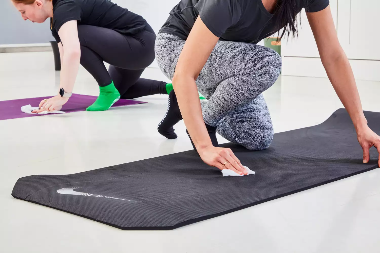 Two women using wipes to clean their yoga mats in a bright fitness studio.