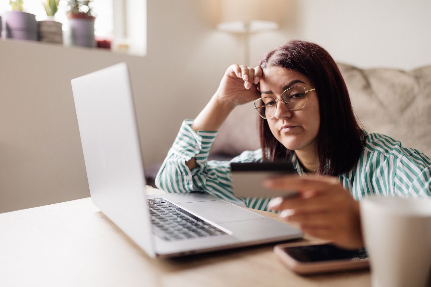 A woman appearing stressed while looking at a credit card and laptop, representing financial anxiety.