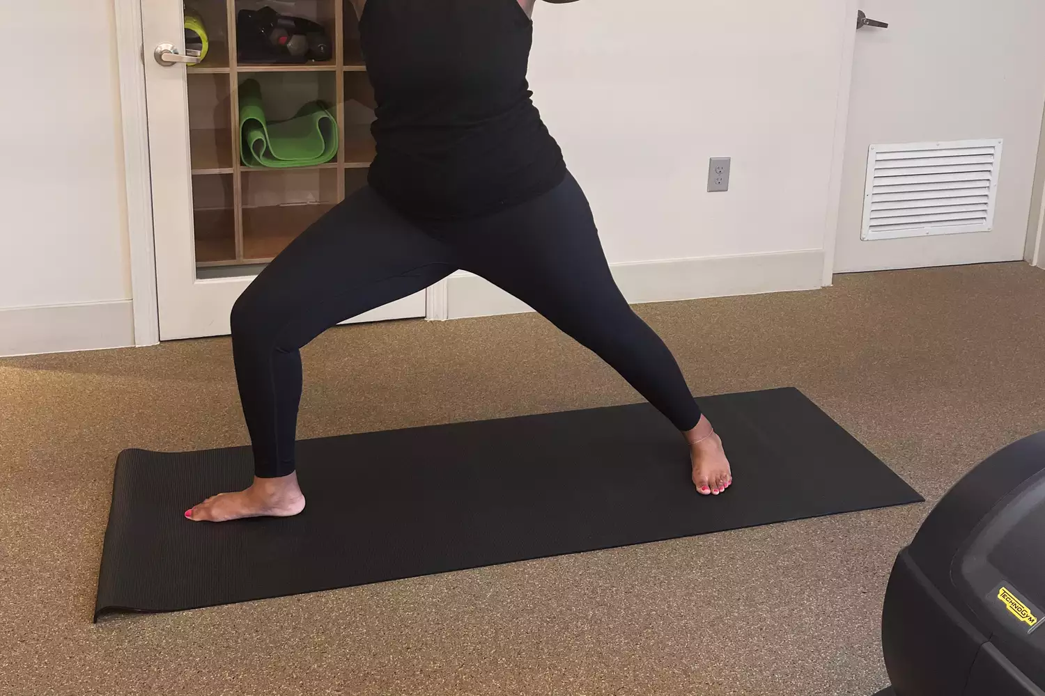 A woman in black athletic wear holding a steady Warrior II yoga pose on a black non-slip mat.
