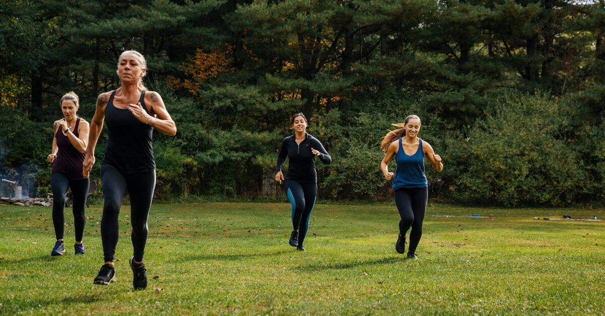 A diverse group of four women wearing athletic gear jogging together in a sunlit park.
