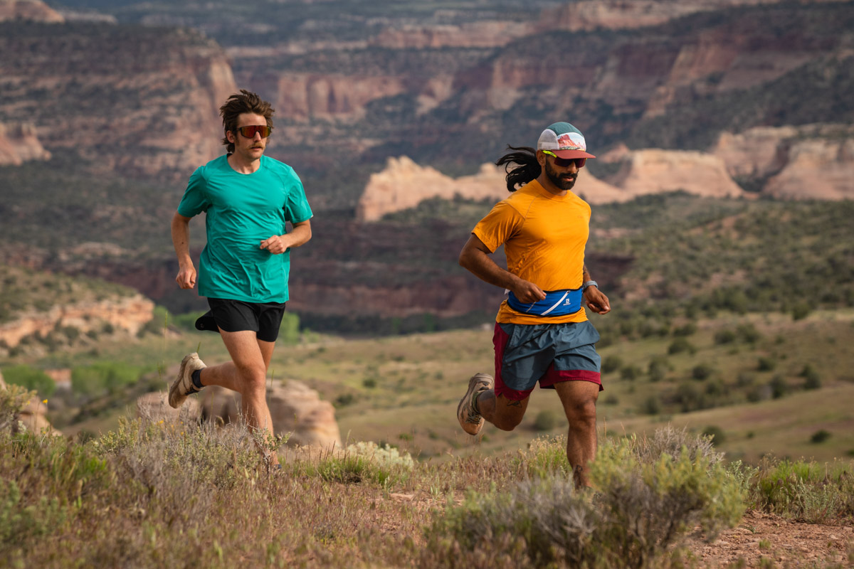 Two runners wearing lightweight gear while running on a sun-drenched desert trail.