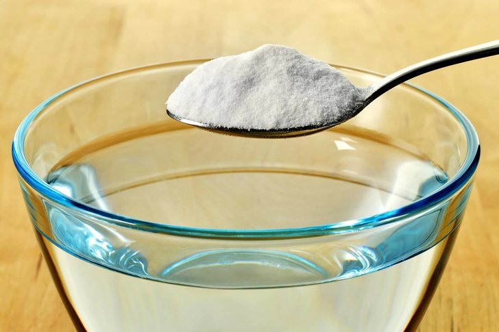 A measuring spoon of white baking soda held next to a glass of clear water.