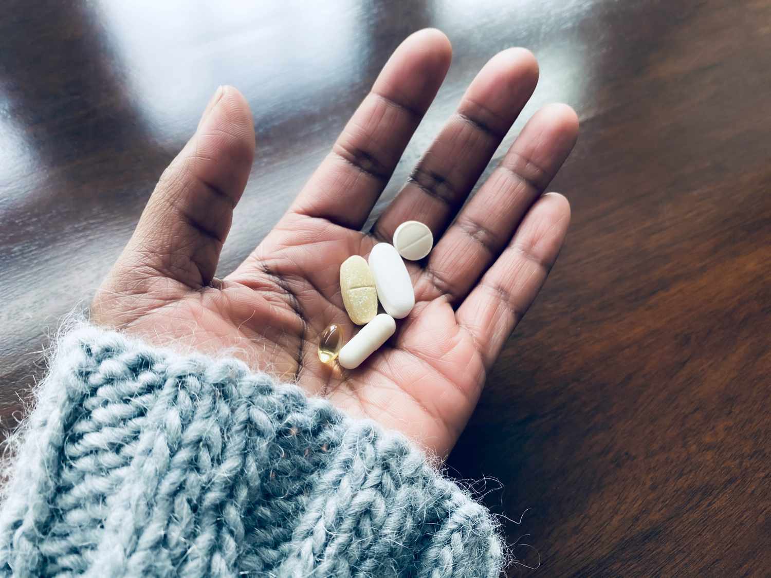 A hand holding a variety of supplement capsules and tablets over a wooden surface.