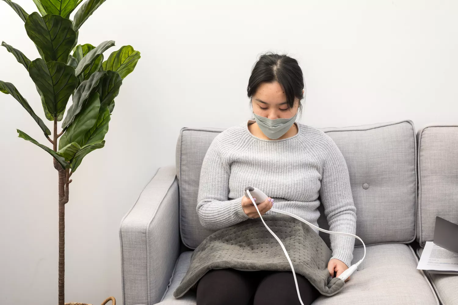 A woman relaxing on a gray sofa using an electric heating pad with a handheld controller.