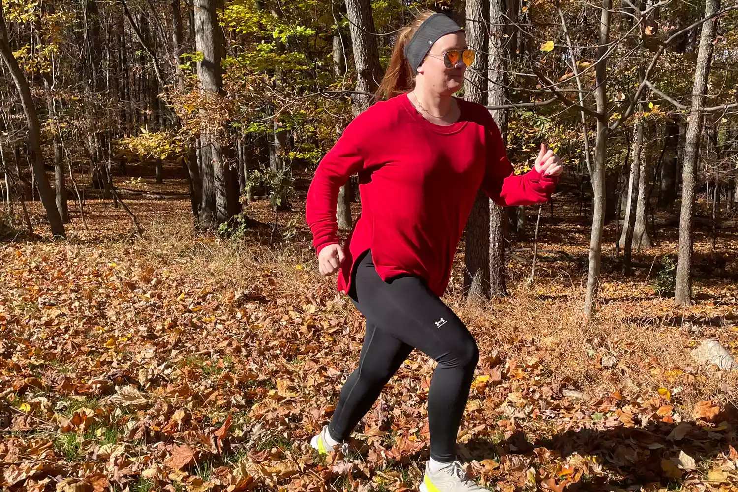 A woman running through an autumn forest trail in black compression leggings and a long-sleeve shirt.