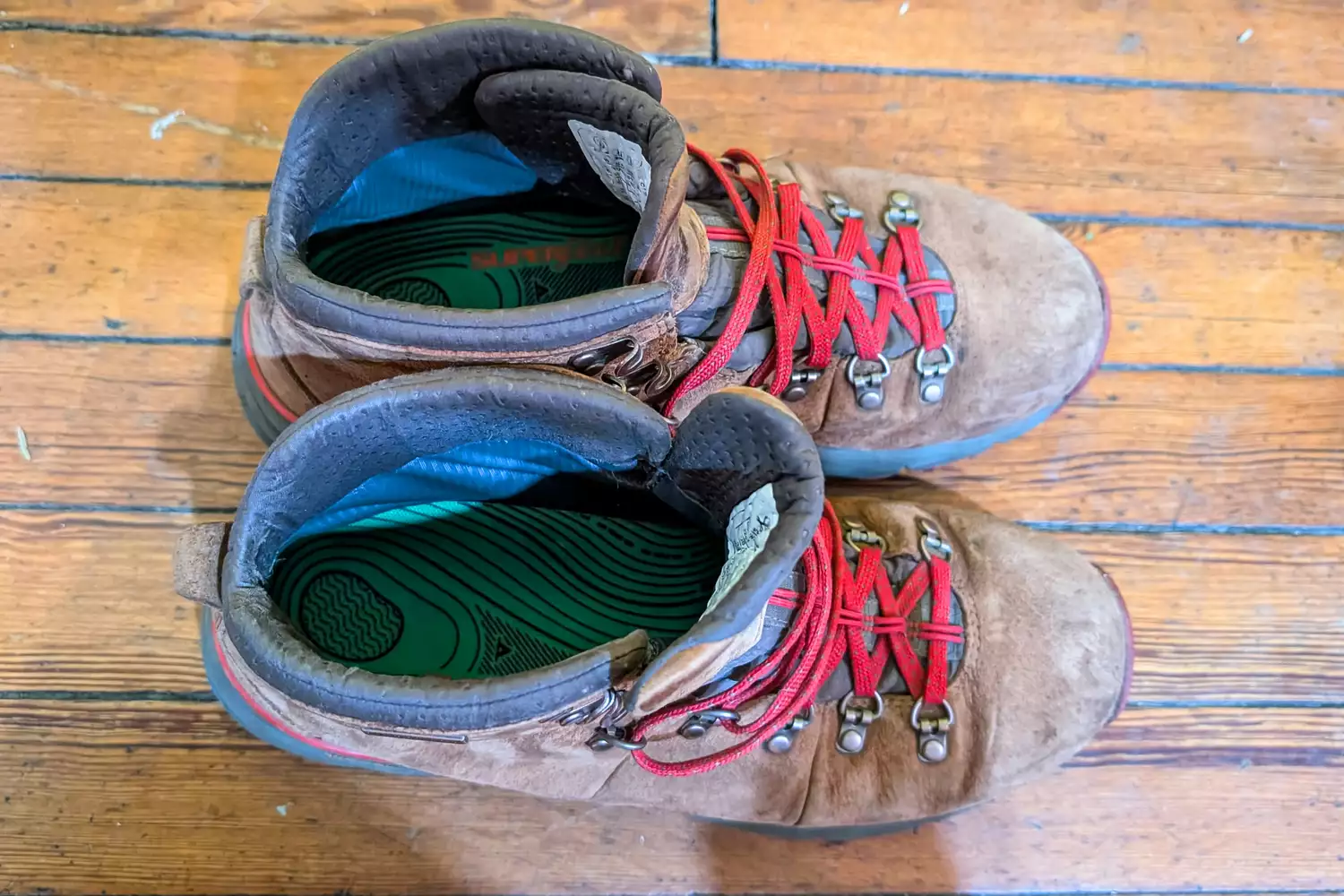 A pair of worn brown leather hiking boots with red laces and green supportive insoles visible inside.