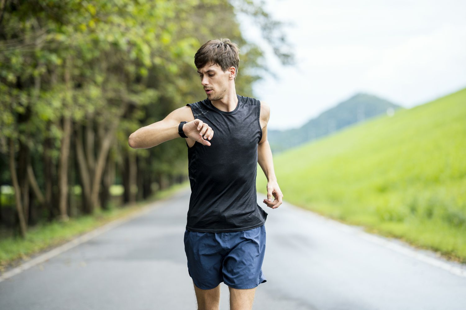 A man jogging outdoors through trees while looking at his smartwatch to monitor his pace and heart rate.