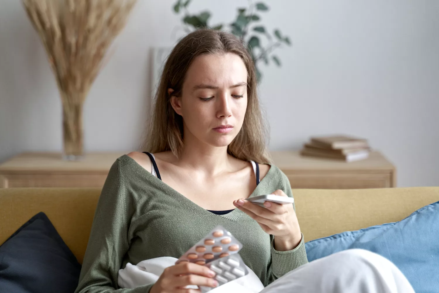 A woman holding supplement pills while checking her phone in a relaxed home setting.