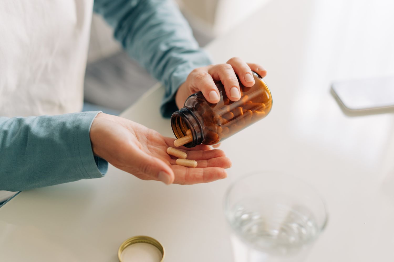Hand pouring vitamin capsules from a bottle next to a glass of water on a table.
