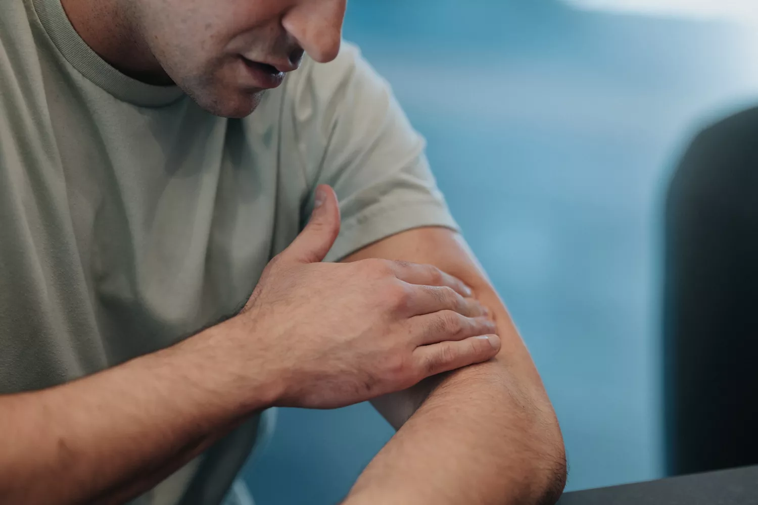 A man in a clinical setting touching his elbow as if checking for soreness or weakness.