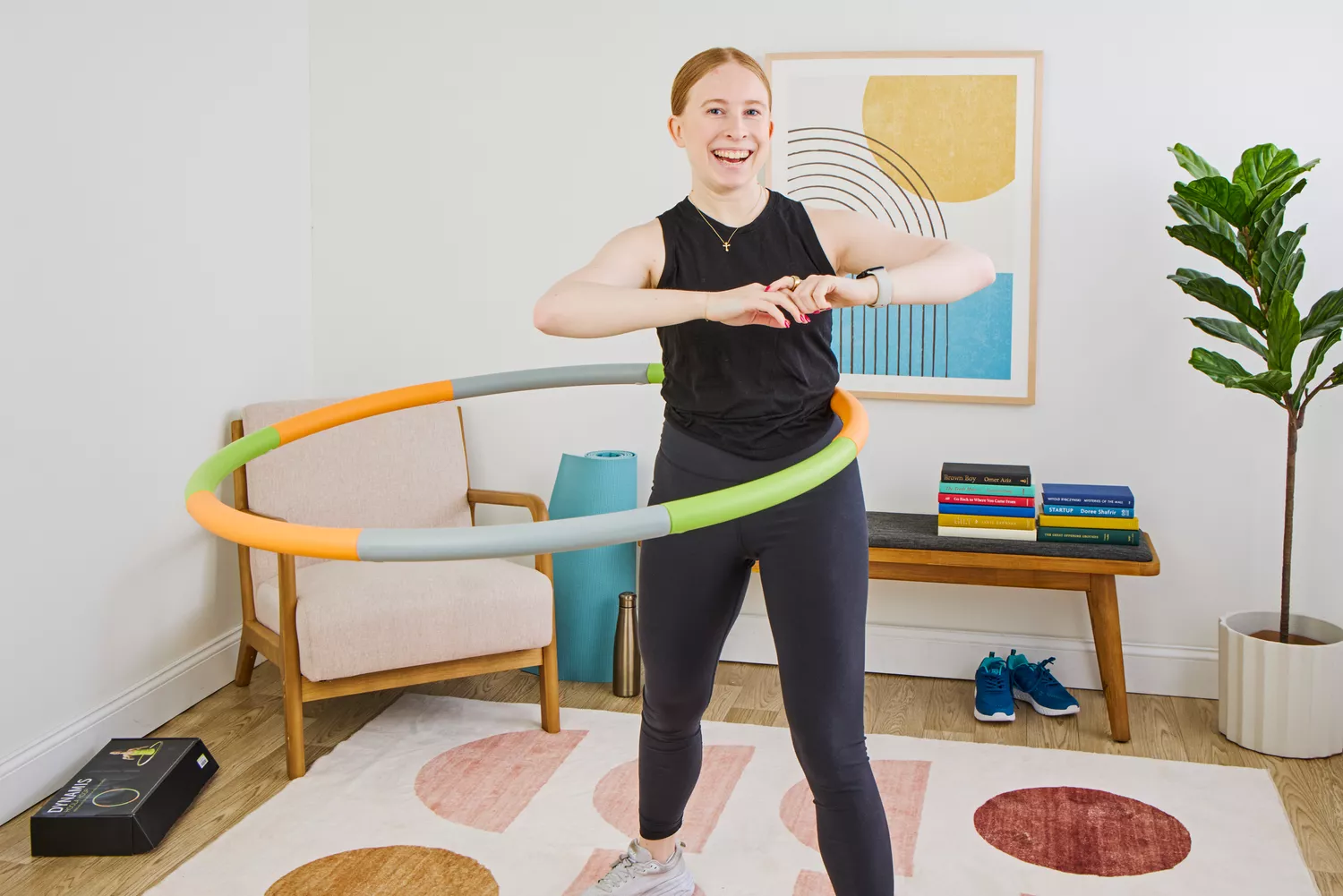 A smiling woman in athletic wear using a multi-colored segmented weighted hula hoop in a bright living room.