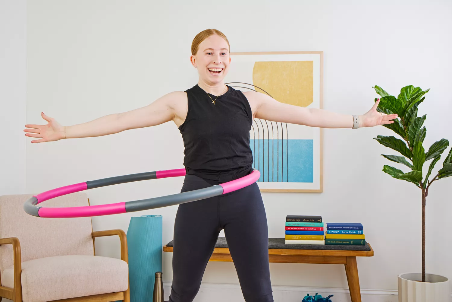 A woman in workout attire performing a core exercise with a pink and gray weighted hula hoop in a modern interior.