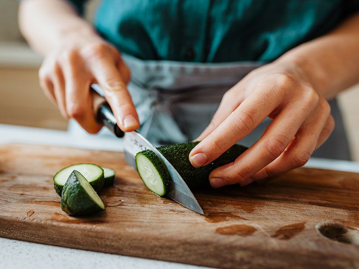 A person slicing a fresh cucumber on a wooden cutting board in a kitchen.