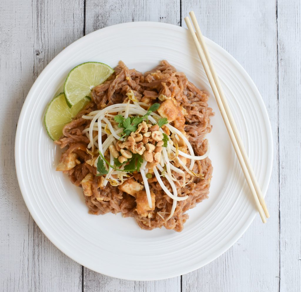 A plate of Pad Thai with rice noodles, peanuts, and lime wedges.