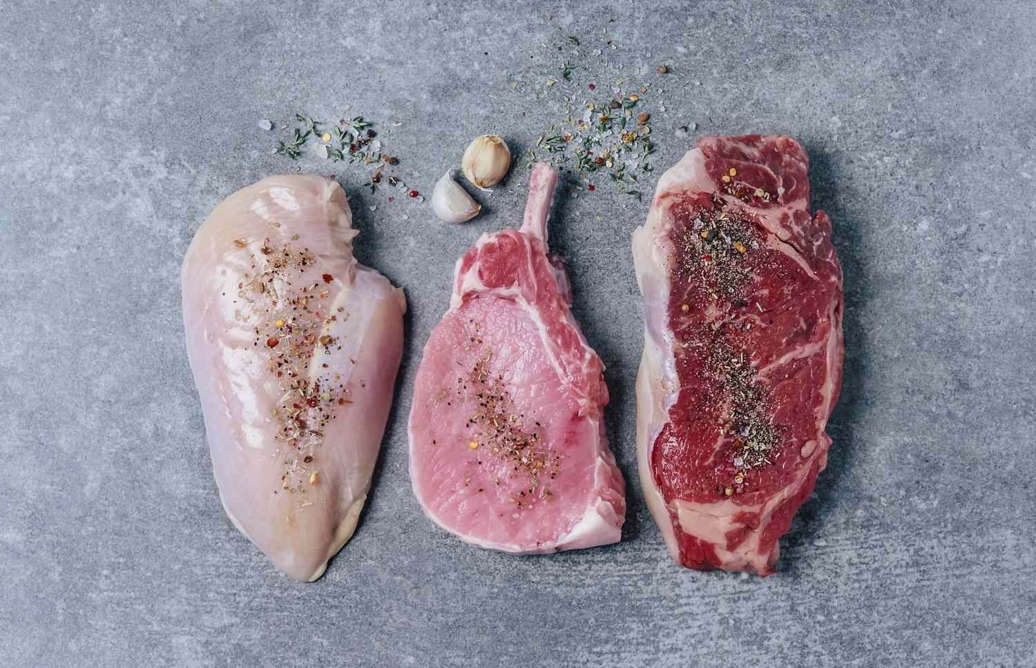 An assortment of raw chicken, pork, and beef steaks arranged on a gray surface with spices.